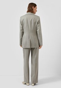 Vrouw met kort bruin haar staat met haar rug naar ons toe, gekleed in een op maat gemaakt lichtgrijs pak met rechte broek en beige hakken.