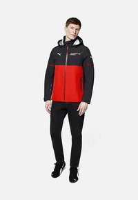Czarna i czerwona wodoodporna kurtka z kapturem, zamek z przodu i akcenty z logo. Stylizowana z czarnymi spodniami i sneakersami.