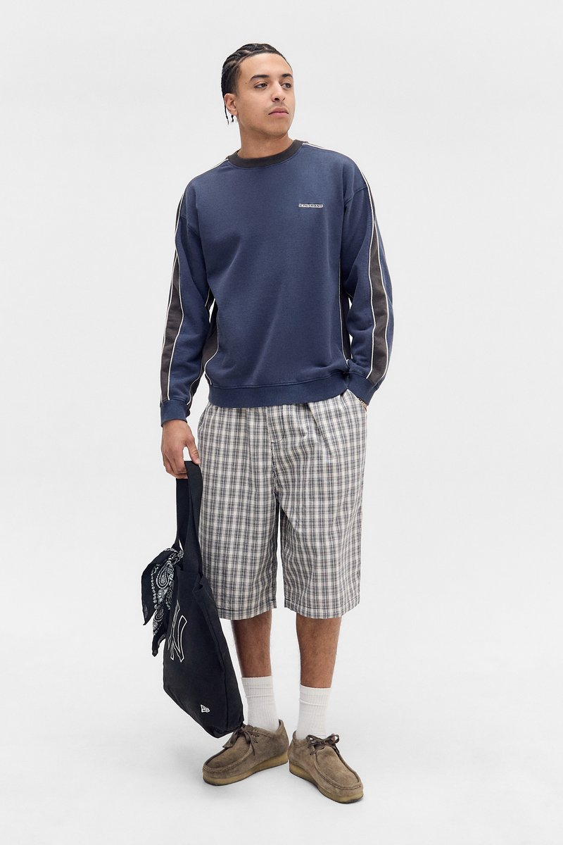 Blå hettegenser med kontrasterende grå sidestropper, sammen med rutete shorts. Brune antikksko i semsket skinn og en svart veske med et bandana-tilbehør.