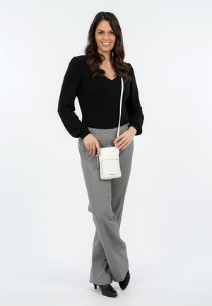 Woman with long dark hair wearing black blouse, gray pants, black heels, and holding small white crossbody bag while smiling.