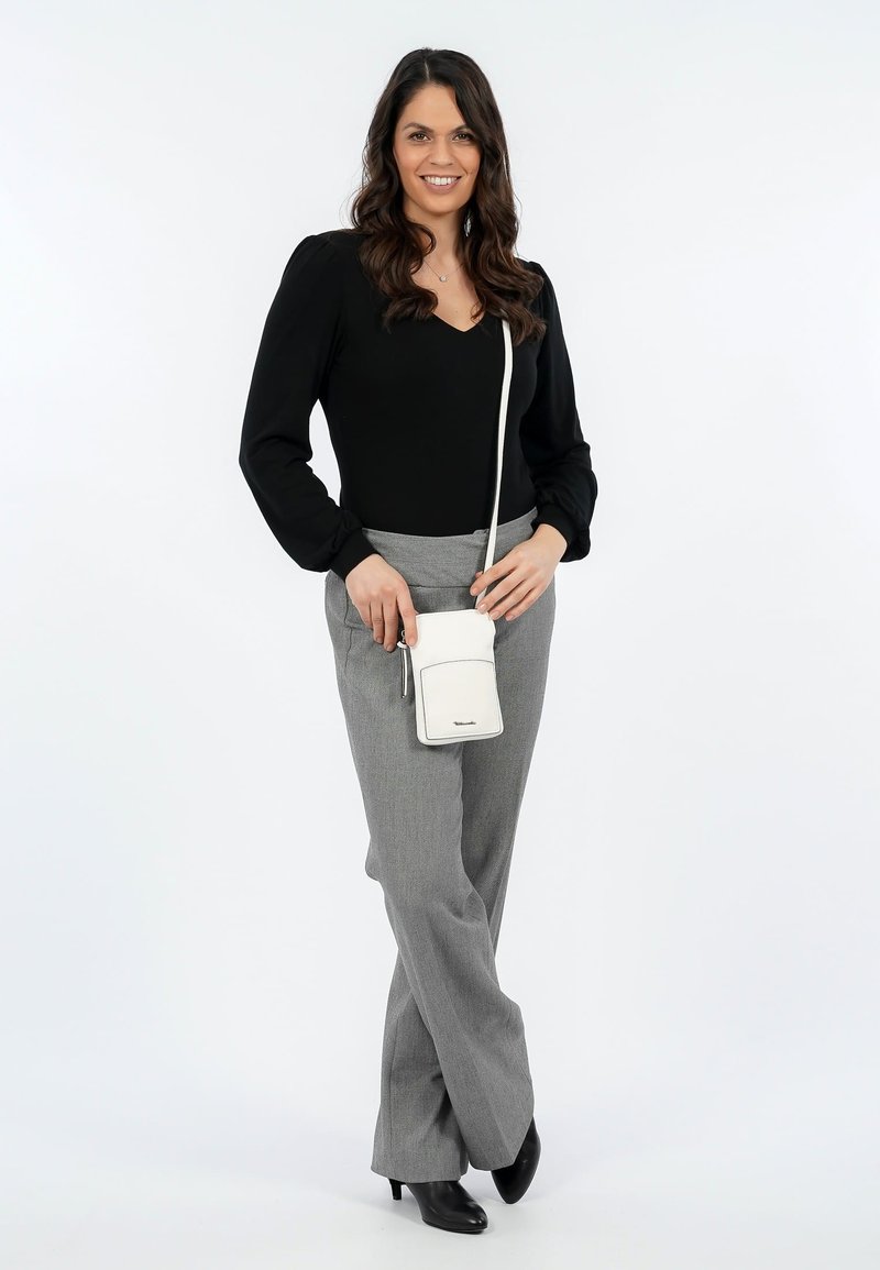 Woman with long dark hair wearing black blouse, gray pants, black heels, and holding small white crossbody bag while smiling.