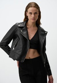 Giacca da biker in pelle nera con accessori in argento, zip asimmetrica e colletto a punta. Il modello indossa un top nero e pantaloni a vita alta.