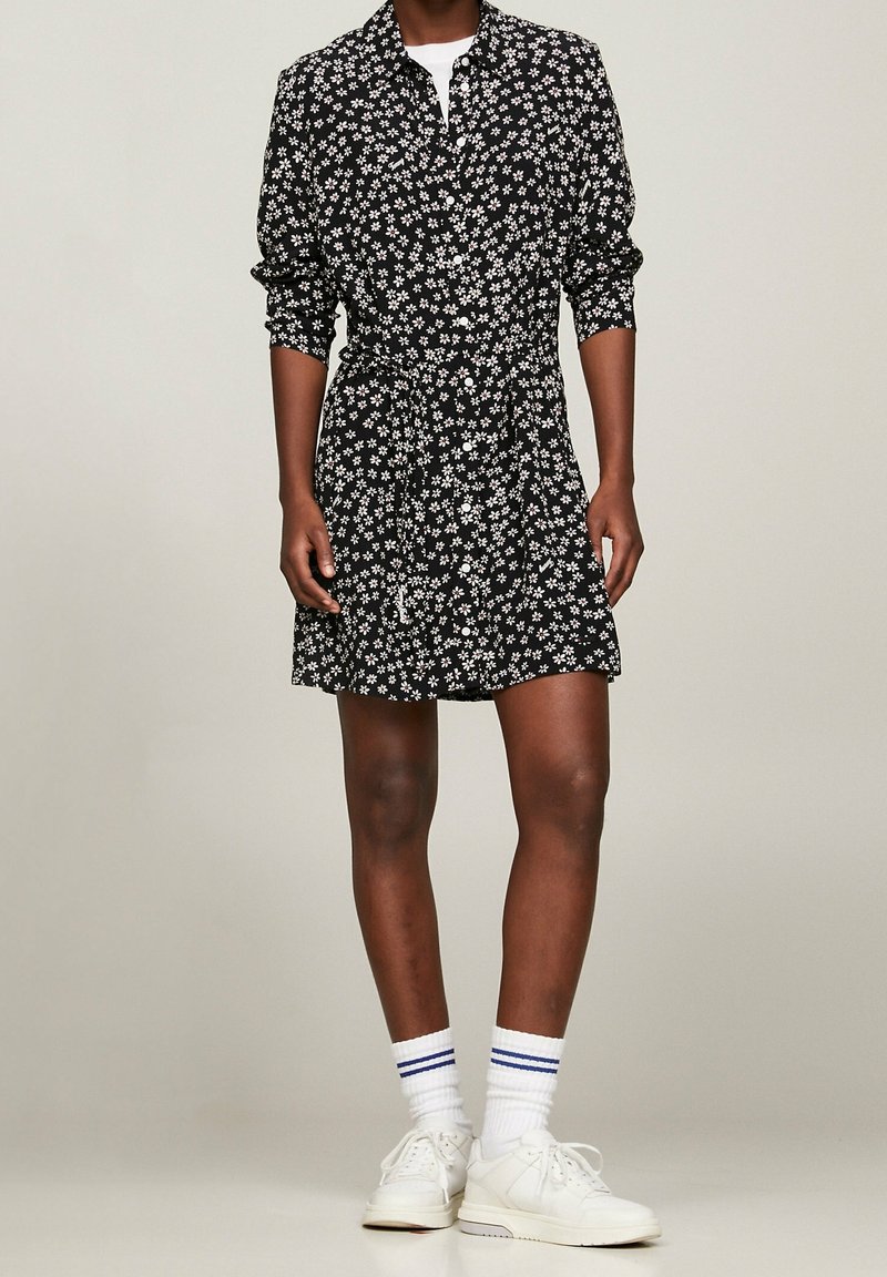 Robe-chemise noire à fleurs avec manches longues et ceinture à la taille ; présente de petites fleurs blanches et une patte de boutonnage sur le devant. Portée avec des baskets blanches.