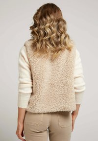 Gilet sans manches beige au texture moelleuse, porté par-dessus un haut à manches longues crème. Le pantalon est de couleur beige clair avec des poches arrière.