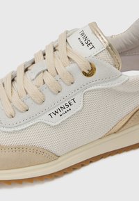 Baskets de sport beiges avec un mélange de mesh et de suède, lacets beiges et accents dorés, étiquette avec logo sur le côté, semelle texturée.