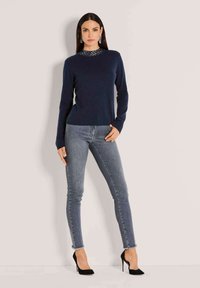 Eine Frau steht an einer hellen Wand und trägt einen marineblauen Pullover, graue Skinny-Jeans mit ausgefransten Säumen, schwarze High Heels und silberne Ohrhänger.
