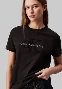 Camiseta de algodón negra con el logotipo bordado de "Calvin Klein Jeans" y "CK" en tejido negro texturizado, con un cuello redondo clásico y mangas cortas.