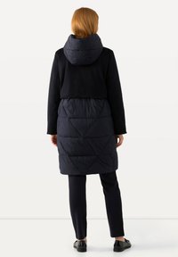 Vadderad jacka med en kortad övre del, marinblå färg, stor huva och geometrisk kviltad design på den nedre halvan. Matchad med mörka byxor och skor.