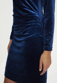 Frau trägt ein dunkelblaues Samtkleid mit langen Ärmeln und gerafftem Detail an der Seite, von der Mitte des Oberschenkels bis zur Taille gezeigt, rechter Arm entspannt an der Seite.
