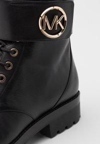 Botín de cuero negro hasta el tobillo con un logotipo dorado redondo en la correa, que presenta un diseño con cordones, ojales dorados y suela de goma negra gruesa.