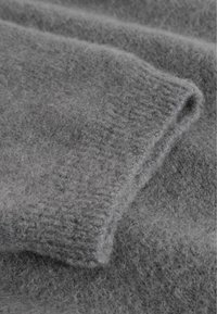 Weicher, strukturierter grauer Strickstoff mit sichtbaren Fasern und einer umgeschlagenen Kante, die auf ein warmes Kleidungsstück aus Wolle oder Mohair hinweist.