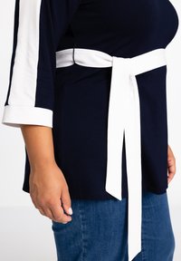 Marineblaues Oberteil mit weißen Akzentstreifen an den Ärmeln und einem weißen Stoffgürtel, der in der Taille gebunden ist. Hat eine weiche, glatte Textur.