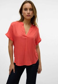 Blusa corallo a maniche corte con scollatura a V. Tessuto morbido e leggero con maniche arrotolate; orlo leggermente curvo per una vestibilità rilassata.