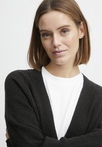 Cardigan noir en tricot avec texture côtelée, design à l'avant ouvert, porté sur un t-shirt blanc. Présente des manches longues et une coupe décontractée.