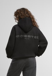 Schwarzer übergroßer Hoodie mit geprägtem Text "VERBOTEN" und "10119, BERLIN" auf dem Rücken. Weicher Stoff, Kapuze mit Kordelzug, lockere Passform.