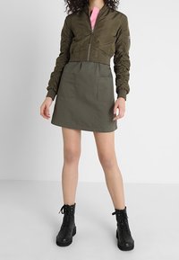 Kobieta w oliwkowozielonej kurtce bombowcu, dopasowanej spódnicy A-line z przednimi kieszeniami, różowej koszuli i czarnych, sznurowanych butach wojskowych stoi na białej podłodze.