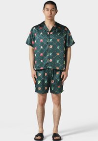 Camisa de seda de manga corta en verde oscuro y pantalones cortos a juego adornados con un patrón floral y geométrico. La textura es suave y brillante.
