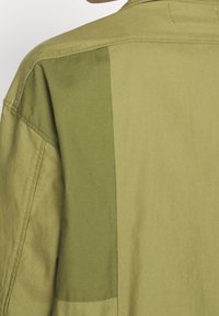 Maglietta verde con un design bicolore composto da sezioni di tessuto verde chiaro e scuro. Il materiale appare liscio e leggero.