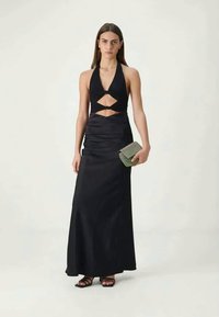 Mulher vestindo um vestido preto de corte halter com recortes, segurando uma pequena bolsa texturizada, em pé sobre um fundo claro e simples.