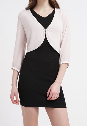 Kobieta nosząca dopasowaną, czarną sukienkę o fakturze, zestawioną z jasnoróżowym krótkim żakietem bolerkiem z rękawami do łokcia.