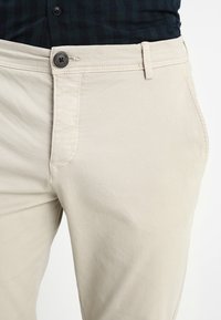 Beige bomullsbyxor med en smal passform, med knappstängning, bälten, och sidofickor, uppvisade mot en neutral bakgrund.