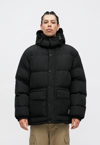 Fekete puffázott dzseki kapucnival, elülső cipzárral és pattintós gombokkal, két elülső zsebbel és rugalmas mandzsettával. Simaságú szövettextúra.