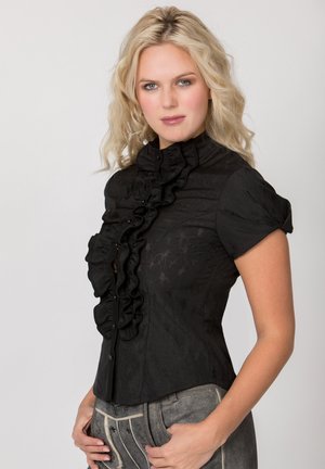 Frau mit blonden, welligen Haaren, die eine taillierte schwarze Bluse mit Rüschen und kurzen Ärmeln trägt, kombiniert mit grauen gemusterten Hosen, steht und schaut nach vorne.