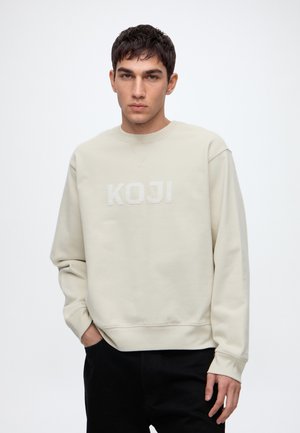 Jeune homme aux cheveux courts et foncés portant un sweat-shirt beige clair avec le texte "KOJI", debout avec une main dans la poche sur fond blanc.