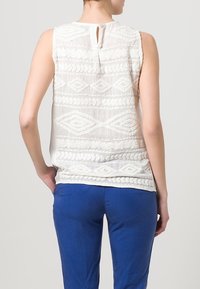 Bílý top bez rukávů s texturovanou výšivkou s geometrickými vzory; se rozparkem na zádech. V kombinaci s modrými kalhotami.