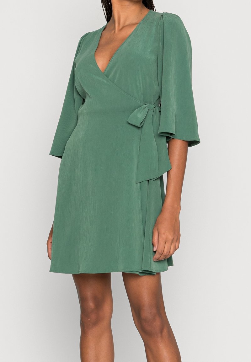Femme portant une robe portefeuille verte mi-longue aux manches trois-quarts avec un lien sur le côté, sur un fond clair uni.