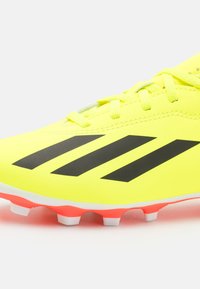 Scarpino da calcio giallo brillante con strisce nere, dotato di una parte superiore in sintetico liscia, chiusura con lacci e suola accentuata di rosa con tacchetti bianchi.
