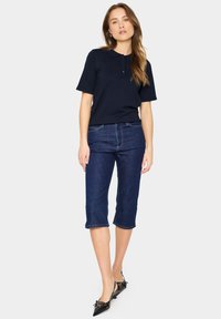 Námořnická polo košile s knoflíky, spárovaná s tmavými denimovými capri kalhotami. Černé boty s dekorativními akcenty dotvářejí celý outfit.