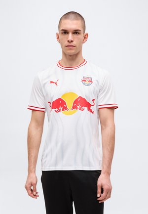 Junger Mann mit kurzen Haaren trägt ein weißes Red Bull Salzburg Fußballtrikot mit roten und gelben Logos und schwarzen Hosen, steht vor einem einfarbigen Hintergrund.