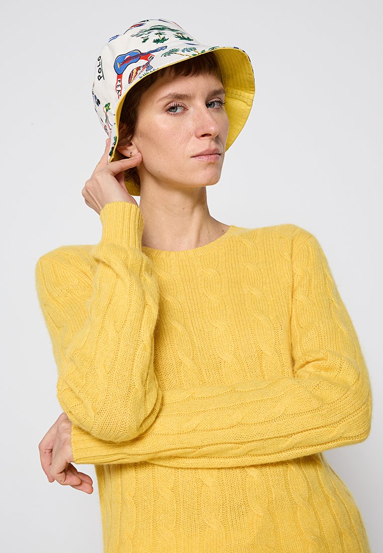 Żółty sweter w sploty wykonany z miękkiej przędzy, zestawiony z białą czapką typu bucket z kolorowymi wzorami gitar i roślin.