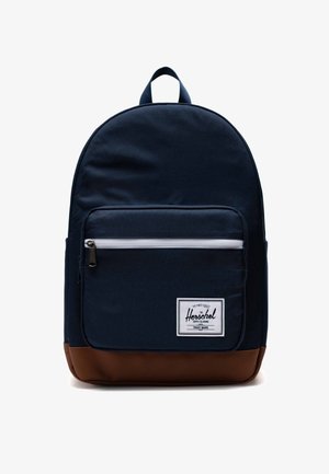 Granatowy plecak materiałowy o gładkiej fakturze, z przednią kieszenią na zamek błyskawiczny, brązowym skórzanym dnem oraz białą łatą z logo.