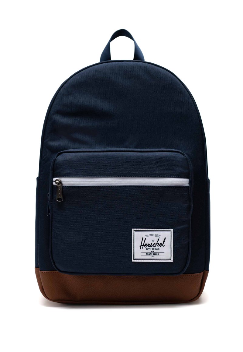 Námornícky modrý látkový batoh s hladkým povrchom, ktorý má prednú zipsovú kapsu, hnedý kožený spodok a bielu nášivku s logom.