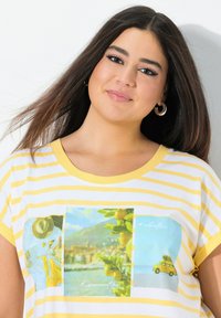 Keltainen ja valkoinen raidallinen t-paita, jossa on kolme värikästä grafiikkaa: aurinkohattu, sitruunapuu ja vintage-auto rannalla. Pehmeää kangasta.
