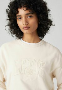 Lichtbeige sweatshirt met een getextureerd LEVI'S-logo geborduurd op de borst. Heeft een ronde hals en zachte stof. Krullend haar zichtbaar.