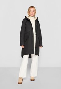 Juodas parka tipo švarkas su užtrauktuku priekyje, kaklaraiščiu reguliuojama gobtuvu ir dviem priekinėmis kišenėmis. Pamuštas smėlio spalvos medžiaga, turi atsipalaidavusį siluetą.