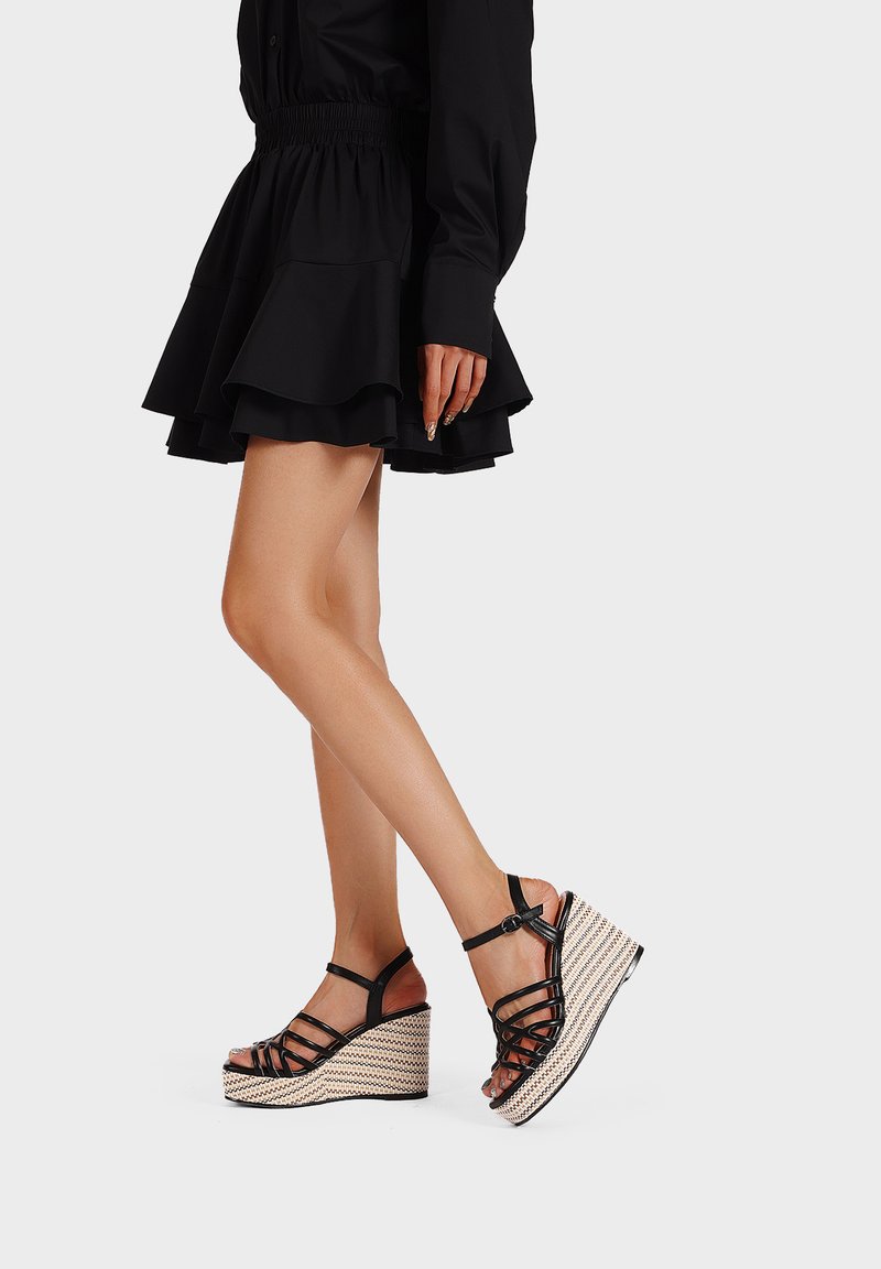 Sandali con zeppa nera e cinghietti, dotati di una suola intrecciata e testurizzata. Presentano un cinturino alla caviglia e un design con punta aperta, evidenziando un'estetica estiva.