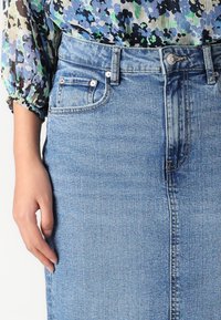 Heledsinine denim seelik, millel on klassikaline kõrge vöökoht, struktureeritud siluett, nööridega detailid ja üks esikott.