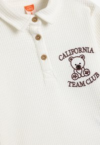 Vit ribbad polo skjorta med en broderad bruns björnlogotyp och texten "CALIFORNIA TEAM CLUB". Tre beige knappar vid kragen.