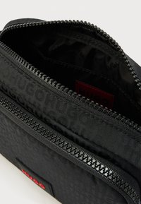 Bustina nera con cerniera realizzata in tessuto testurizzato con un motivo a logo ripetuto, etichetta interna rossa e dettagli metallici eleganti.