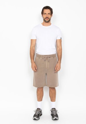 Mann steht frontal und trägt ein weißes T-Shirt, beige Shorts mit Kordelzug, weiße Socken und schwarze Sportschuhe vor einem schlichten weißen Hintergrund.