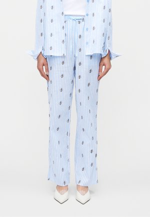 Pantalon à rayures verticales bleu et blanc avec motifs d'abeilles brodés, porté avec une chemise assortie et des chaussures blanches à bouts pointus sur un fond uni.