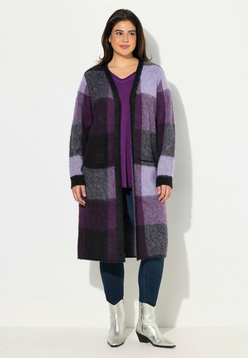 Langer Cardigan im lila und schwarzen Karo-Muster, aus strukturiertem Wolle. Offenes Frontdesign, kombiniert mit einem lila Oberteil und blauen Jeans. Silbergraue Ankle-Boots.