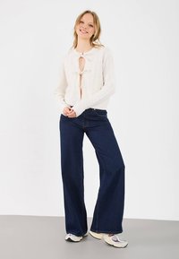 Witte gebreide cardigan met een strik aan de voorkant, kabelgebreide textuur, gecombineerd met donkerblauwe wijde jeans en crèmekleurige sneakers.