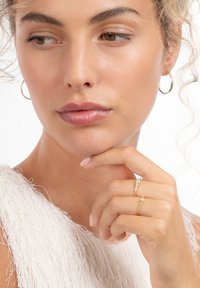 Woman with curly hair wearing gold hoop earrings, a white textured top, and two gold rings with small diamonds, resting hand near chin.
