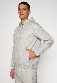 Chaqueta gris claro con capucha, cierre de cremallera, panel superior texturizado con patrón de diamante, puños elásticos y bolsillos laterales.