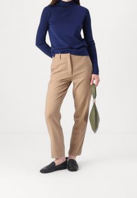 Femme portant un col roulé bleu marine, un pantalon beige, des mocassins noirs, tenant un sac à main vert olive par le poignet d'une main.
